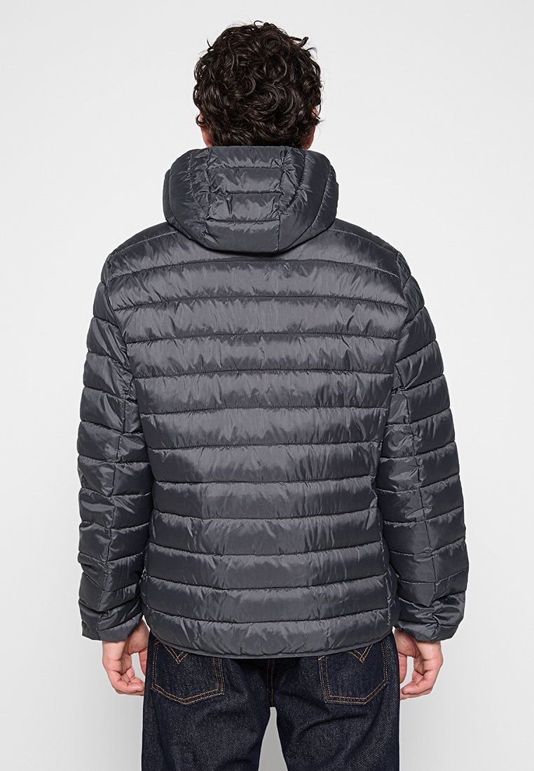 Giacca puffer grigia con cappuccio, caratterizzata da linee quiltate orizzontali, texture liscia e polsini elastici. Indossata sopra jeans di denim scuri.