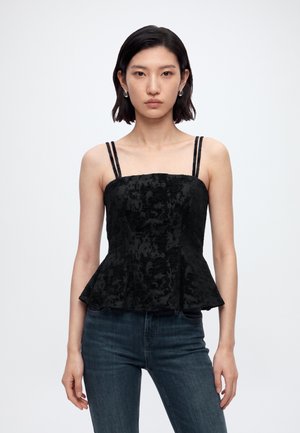 Jeune femme aux cheveux courts et foncés portant un haut noir sans manches à double bretelles et un jean bleu foncé sur fond blanc.