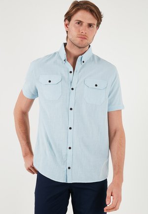 Camisa de manga corta en azul claro, hecha de tejido texturizado, con dos bolsillos frontales con solapas, botones negros y un cuello clásico.