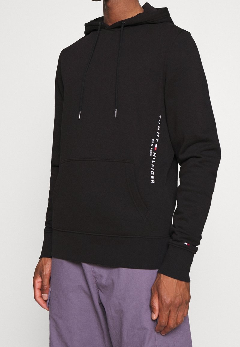 Sudadera con capucha negra con bolsillo frontal y texto "Tommy Hilfiger" en el lateral, combinada con pantalones morados, se muestra desde el cuello hasta las caderas.