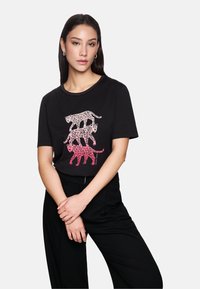 Woman wearing black t-shirt with three stacked pink leopard prints, paired with black pants, standing with hands loosely clasped.