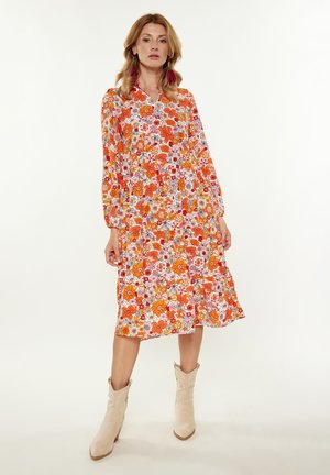 Blumiges Midikleid mit weißem Hintergrund und orangefarbenen, roten und grauen Blumen, langen Ärmeln und einer Knopfleiste. Kombiniert mit beigen Ankle Boots.
