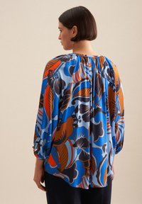 Blusa fluida blu, con un motivo floreale arancione e marrone. Maniche lunghe con polsini arricciati e scollatura arrotondata. Texture liscia.
