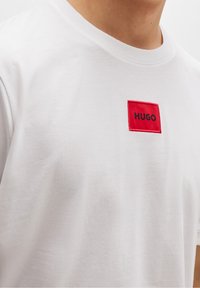 Camiseta de algodón blanca con un parche rectangular rojo que lleva la palabra "HUGO" en bordado negro. Diseño clásico de cuello redondo.