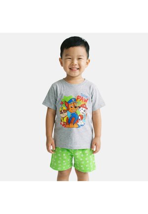 Jeune garçon souriant portant un t-shirt gris avec des personnages de Pat' Patrouille et un short vert vif avec des motifs floraux blancs sur un fond uni.