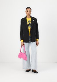 Blazer nero con accessori in argento, maglione grafico giallo, jeans a zampa d'elefante blu chiaro, borsa rosa e ballerine nere. Texture lisce in tutto l'outfit.