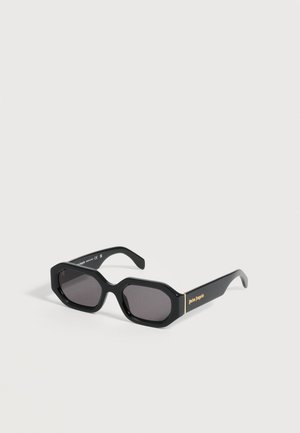 Lunettes de soleil hexagonales noires avec verres foncés et logo doré "palm angels" sur la branche, présentées sur un fond clair neutre.