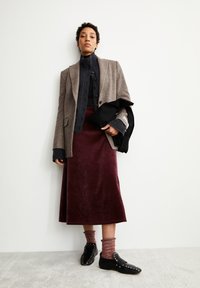 Bruine wollen blazer over een donkere gebreide top, gecombineerd met een kastanjebruine fluwelen rok, rood en grijs gestreepte sokken en zwarte schoenen met studs.