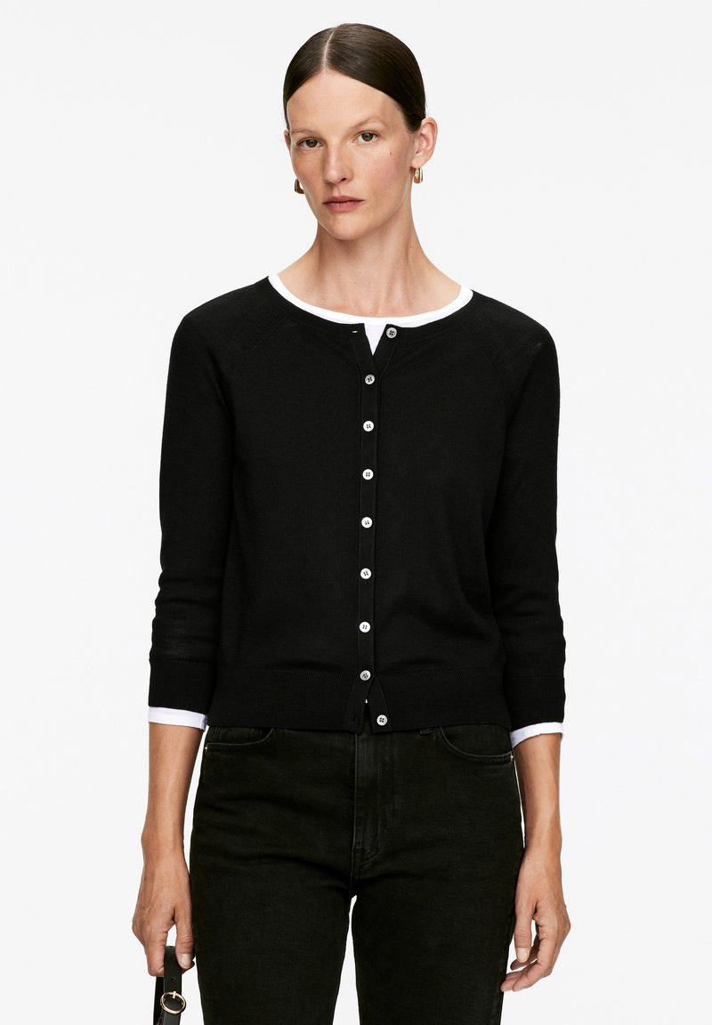 Cardigan noir en tricot doux, avec un col rond, des bordures blanches et une fermeture à boutons. Manches longues avec des poignets côtelés.