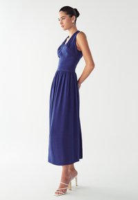 Maksi haljina od tamnoplave tkanine s uskim korzetom, dubokim V-izrezom i skupljenim strukom, koja prelazi u ravni rub. Model nosi prozirne blok potpetice.
