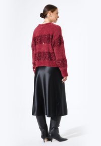 Suéter de punto rojo con rayas de lentejuelas alternas, combinado con una falda midi de satén negra y botas de tacón puntiagudas negras hasta la rodilla.