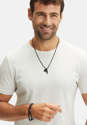 Schwarzes Lederhalsband mit strukturiertem Anhänger, über einem schlichten weißen Hemd getragen. Schwarzes geflochtenes Armband am Handgelenk, das die kontrastierenden Materialien hervorhebt.