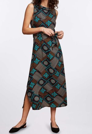 Femme ajustant une robe midi sans manches à motifs avec des fleurs turquoise et des formes géométriques, portant des chaussures plates noires sur fond blanc.