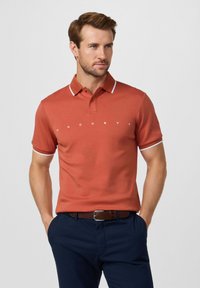 Polo arancione con colletto e dettagli delle maniche bianchi. Presenta un logo bianco sul petto. Indossata con pantaloni scuri e una cintura marrone.