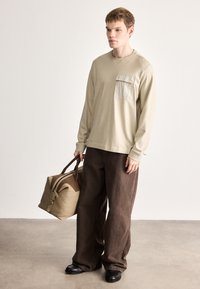 Béžová košeľa s dlhým rukávom a náprsné vrecko; hnedé široké nohavice; drží svetlohnedú cestovnú tašku s hnedými detailmi a zipsom. Čierna obuv.