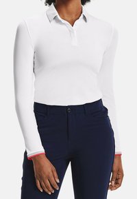 Polo blanc à manches longues avec col, fermeture par boutons et poignets soulignés de rouge ; porté avec un pantalon bleu marine.