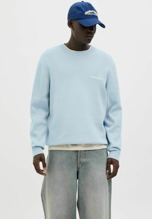 Light blue sweatshirt with ribbed cuffs, featuring a small white logo. Paired with faded denim pants and a blue cap. Smooth texture.