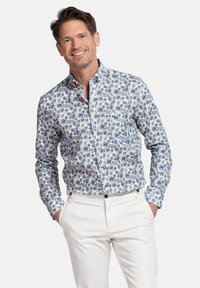Chemise boutonnée en coton blanc avec un motif floral bleu et vert, manches longues et col classique, associée à un pantalon blanc.