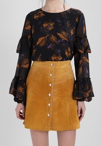 Blouse noire à motifs floraux avec manches volantées associée à une jupe en daim jaune moutarde, dotée d’une fermeture boutonnée frontale et de coutures diagonales.