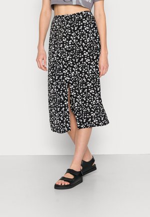 Jupe midi noire à fleurs blanches, avec une fente, en tissu léger, associée à des sandales noires à brides épaisses et semelle plate.