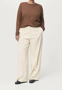 Pull marron à manches longues associé à un pantalon large couleur crème. Le pull est en matériau doux ; le pantalon comporte des plis à l'avant et des poches latérales.