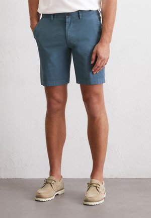 Lichtblauwe shorts met een vlakke voorkant, zijzakken en opgerolde zoom, gecombineerd met beige suède schoenen met witte zolen en veters.