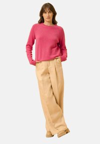 Maglione rosa a maglia con scollo rotondo e polsini a coste, abbinato a pantaloni beige a gamba larga. La modella sta in piedi con le mani in tasca.