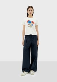 Witte t-shirt met een blauw en rood bloemenprint, gecombineerd met loszittende marineblauwe broeken en witte sneakers met beige accenten.