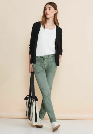 Black long cardigan over a white top, green pants with a relaxed fit, and a striped black-and-white tote bag. White sneakers complete the look.