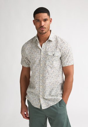Chemise à manches courtes en tissu léger, présentant un motif floral bleu et jaune sur fond blanc, avec une poche poitrine et des boutons.