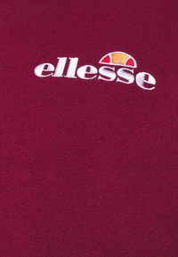 Burgunderfarbenes Gewebe mit weißem, gesticktem "ellesse"-Logo und halbkreisförmigem Design in Orange und Rot über dem Text.