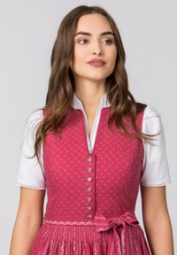 Rosa besticktes Kleid mit tailliertem Oberteil, weißer Bluse darunter, dekorativen Knöpfen und gebundenem Taillengürtel. Zarte Spitzelemente.