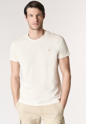 Uomo con capelli scuri in piedi di fronte, indossa una maglietta tinta unita bianco sporco e pantaloncini cargo beige chiaro, mani nelle tasche, sfondo neutro.