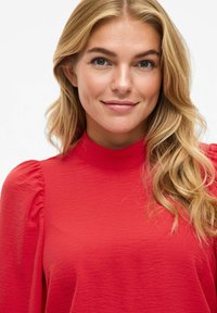 Blusa rossa con texture, collo alto e maniche a sbuffo. Tessuto liscio con sottili pattern irregolari. Inquadratura ravvicinata che mette in evidenza i dettagli.