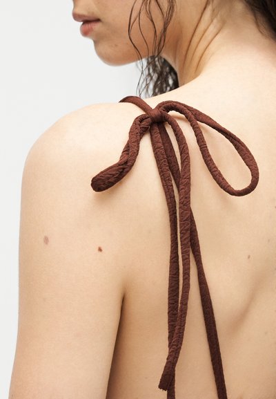 Gros plan sur l'épaule nue et le haut du dos d'une femme avec une bretelle en tissu marron texturé nouée en un nœud papillon.