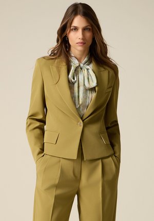 Kvinde iført olivengrøn skræddersyet blazer og matchende bukser med blomstret bluse bundet ved halsen, stående med hænderne i lommerne.