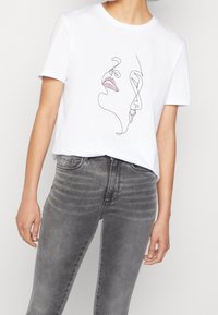 Vit t-shirt med svart kontinuerlig linjekonst av två ansikten, ett med röda läppar, bärs med högmidjade blekta grå jeans.