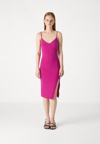 Robe midi rose ajustée avec des fines bretelles, présentant un design sans couture et une fente sur le côté. Accompagnée de sandales noires à lanières.