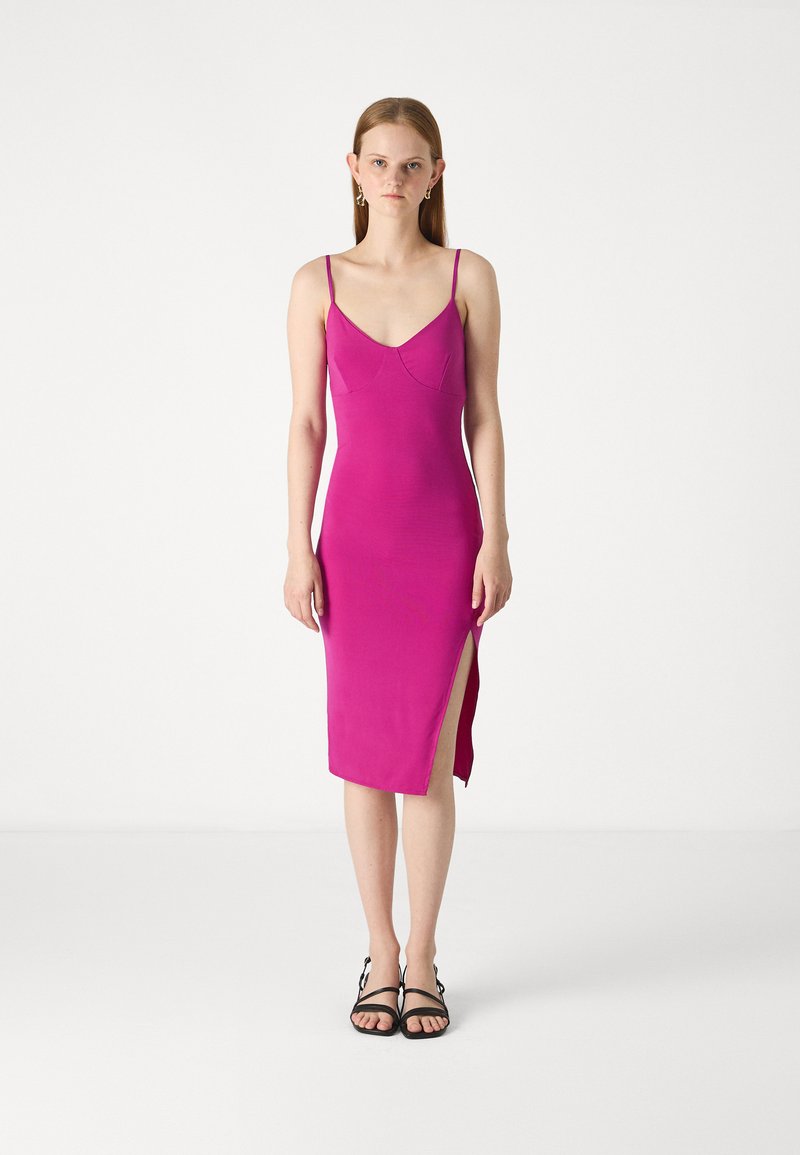 Robe midi rose ajustée avec des fines bretelles, présentant un design sans couture et une fente sur le côté. Accompagnée de sandales noires à lanières.