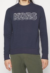 Námořnický modrý svetr s dlouhými rukávy, žebrovanými manžetami a lemem. Na přední straně má bíle orámovanou logo "KORS". Měkká textilie.