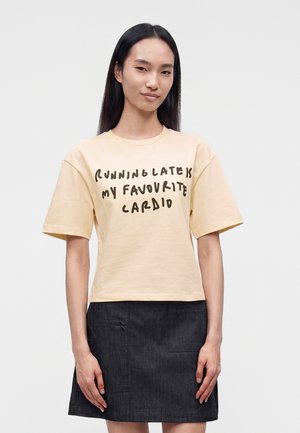 Femeie cu păr drept și închis la culoare poartă un tricou bej pe care scrie "Running late is my favourite cardio" și o fustă închisă la culoare, stând cu fața înainte.