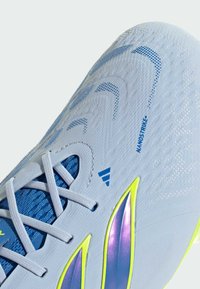 Gros plan d'une chaussure de football Adidas blanche avec des lacets bleus, un tissu bleu texturé, des rayures irisées violettes et jaunes, et le texte "NANOSTRIKE+".