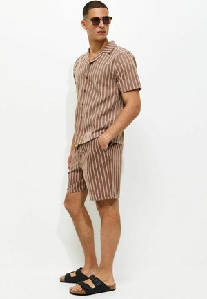 Mężczyzna noszący brązową i białą koszulę w pionowe paski z krótkim rękawem oraz pasujące krótkie spodenki, czarne sandały i ciemne okulary przeciwsłoneczne, stojący z rękami w kieszeniach.