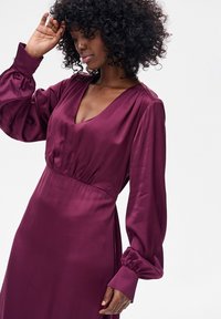 Bordowa satynowa sukienka z głębokim dekoltem w serek, marszczonym talią i długimi bufkami. Gładka tekstura z delikatnym połyskiem i minimalistyczny design.