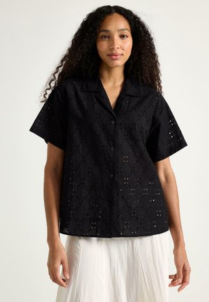 Kvinde med langt, krøllet hår iført en kortærmet, sort øjet knap-up skjorte og en hvid nederdel, stående foran en ensfarvet baggrund.