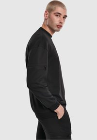 Felpa nera realizzata in tessuto morbido, con collo tondo, maniche lunghe, tasca laterale con zip e polsini e orlo a costine.
