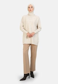 Vaalean beige neulepaita, jossa on sukkulakuvio, pitkät hihat ja korkea kaulus, yhdistettynä istuviin beigeihin housuihin ja teräväkärkisiin mustiin korkokenkiin.