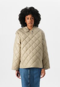 Veste matelassée beige avec col, présentant un motif en losanges, coupe décontractée et manches longues. Portée par-dessus un jean bleu.