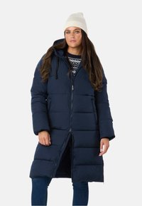 Marineblå dunjakke med hætte, lynlås foran og store lommer. Har et quiltet design og blød tekstur. Båret sammen med en mønstret sweater.