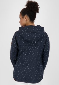 Giacca blu navy con cappuccio, caratterizzata da un motivo di cerchi blu chiaro. Realizzata in tessuto liscio con una vestibilità morbida e dettaglio della yoke sul retro.